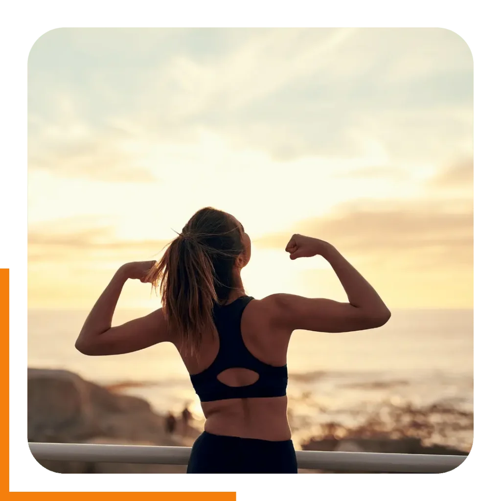 A young woman triumphantly flexing her arms at sunset, reflecting the positive results of online personal training.